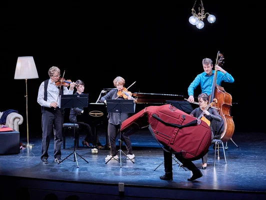 Bühne mit Musiker*innen, ein Musiker versteckt sich in einer Cello-Hülle